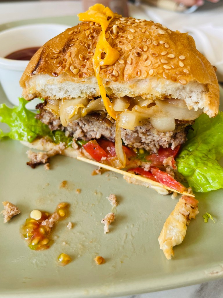 A close-up of a partially eaten The Coffee Library Cheese Burger on a green plate, featuring a sesame seed bun, lettuce, tomato, onions, and melted cheese, accompanied by a small bowl of sauce.