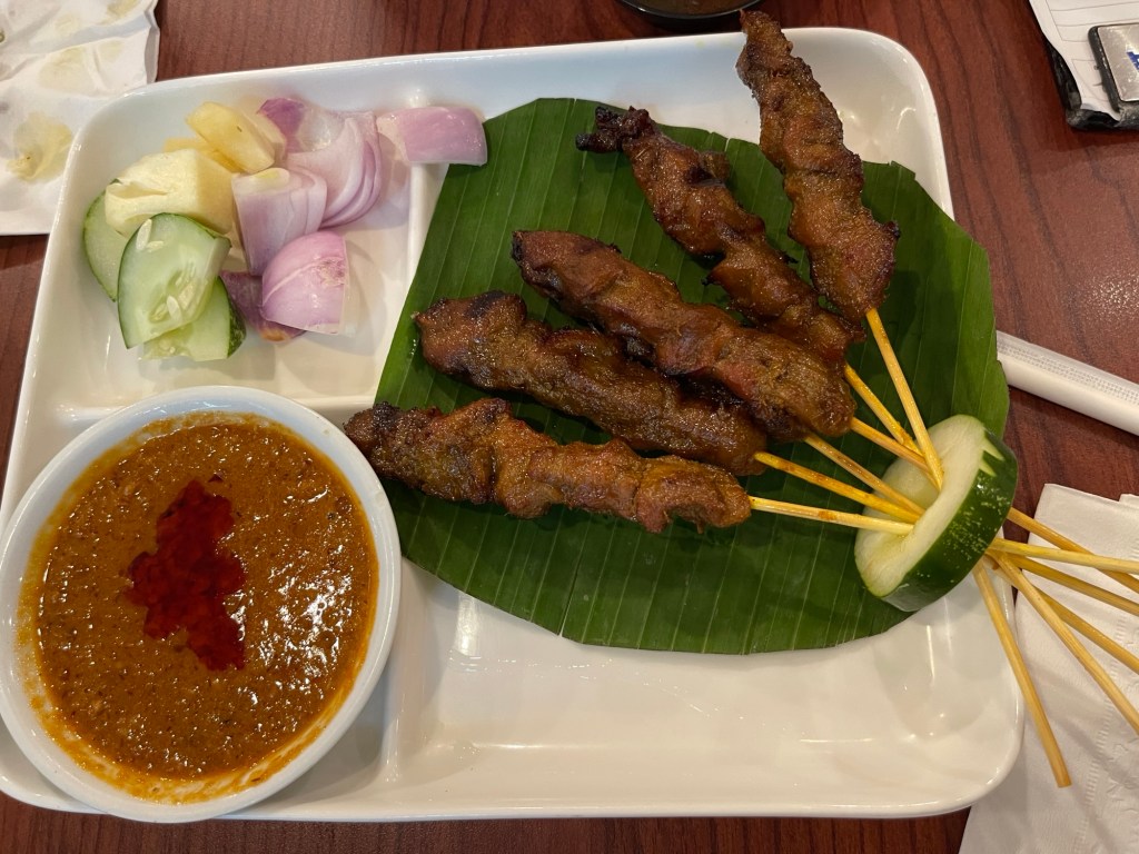 1 order of Chicken Satay with peanut sauce and side dishes: slices of cucumber, pineapple and red onions